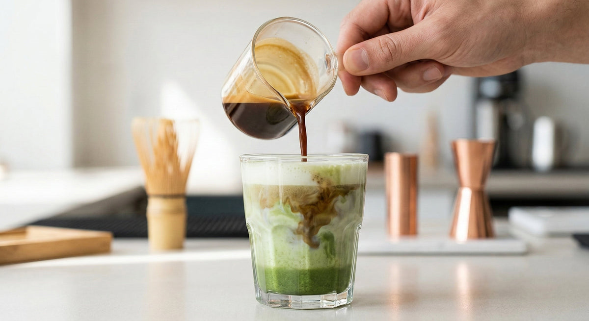 Espresso shot poured into a glass of iced matcha latte creating layered swirls on a kitchen counter with copper tools