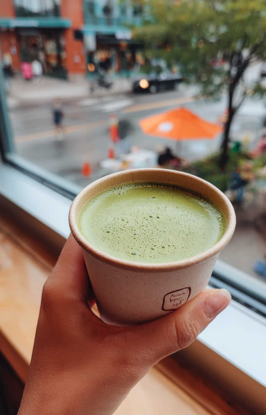 Hand holding a paper cup of hot matcha latte by a window overlooking a city street, cozy cafe takeaway moment