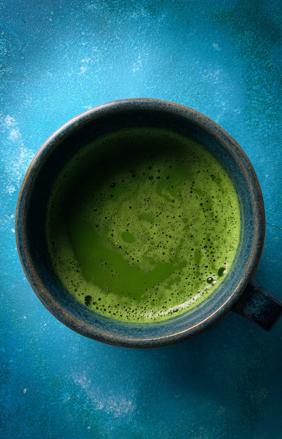 Dark ceramic bowl of frothy matcha on a textured blue surface, overhead shot with strong light and shadow contrast