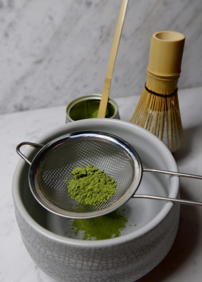 Matcha green tea powder in a small bowl with a whisk and spoon on a marble surface.
